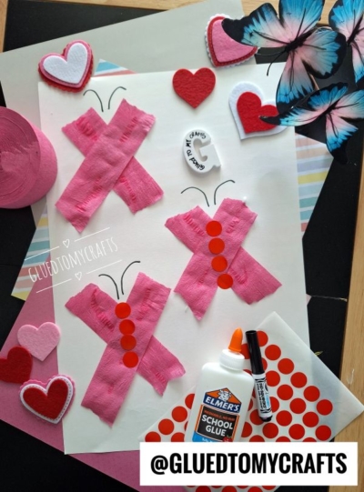 A craft project with four butterflies made from pink tissue paper Xs, red dot stickers for bodies, and marker antennae on white paper. Surrounding are felt hearts, a glue bottle, marker, paper, and butterfly cutouts.