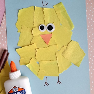 A chick craft made from torn yellow paper pieces glued on blue paper, with googly eyes and an orange beak. A bottle of Elmer’s glue and a brown crayon are nearby. @GLUEDTOMYCRAFTS is written at the bottom.