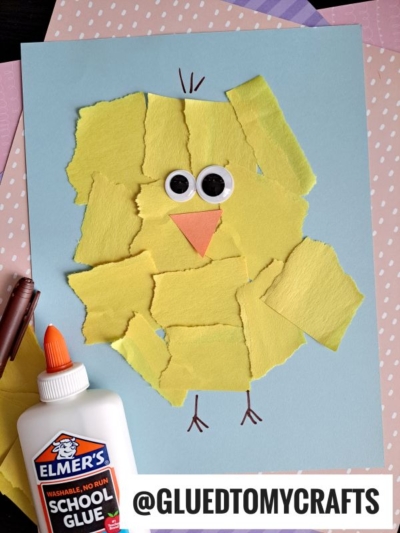 A chick craft made from torn yellow paper pieces glued on blue paper, with googly eyes and an orange beak. A bottle of Elmer’s glue and a brown crayon are nearby. @GLUEDTOMYCRAFTS is written at the bottom.