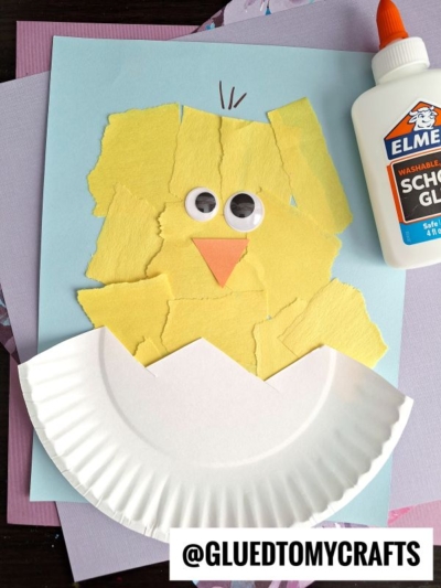 A craft of a chick made from torn yellow paper pieces, large googly eyes, and an orange paper beak on blue paper. A paper plate forms a cracked eggshell at the bottom. Elmers glue is on the right.