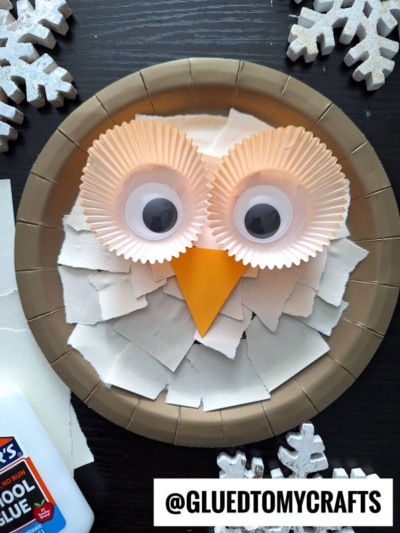A paper plate owl craft features white torn paper, cupcake liners for eyes, large googly eyes, an orange paper beak, and snowflake decorations on a dark surface. A bottle of glue is nearby.