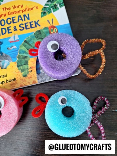 Colorful fish crafts made from pool noodles, pipe cleaners, and googly eyes are displayed on a dark surface next to a children’s “Ocean Hide & Seek” book. The @gluedtomycrafts handle is visible at the bottom.