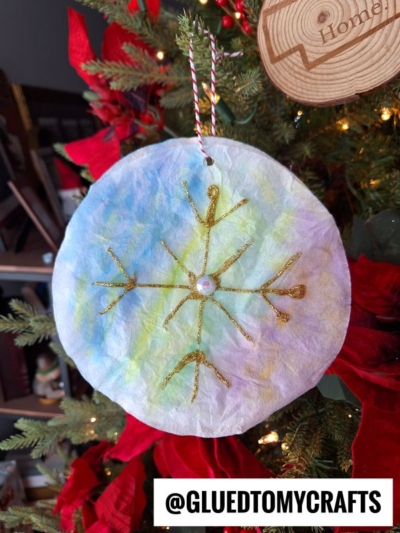 A round, tie-dyed coffee filter ornament with a gold glitter snowflake and a pearl center hangs on a Christmas tree. The background features red poinsettias. Text at the bottom reads @GLUEDTOMYCRAFTS.