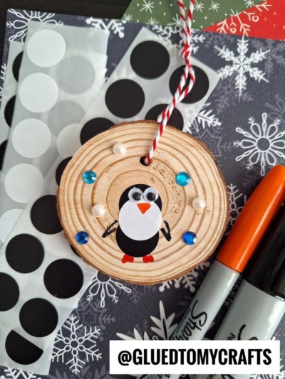 A wooden ornament with a penguin made from paper shapes, coding label crafts, and decorated with sequins and pearls lies on snowflake-patterned paper next to black and orange Sharpie markers and a sheet of black dot stickers.