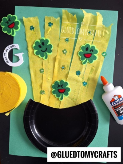 A St. Patrick’s Day craft featuring a paper plate leprechaun cauldron, yellow crepe strips, felt shamrocks with smiley faces, green confetti, glue, a craft knife, and yellow felt on green paper.
