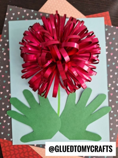 A handmade flower craft with a red, curly ribbon flower and two green handprint cutouts as leaves, arranged on colored paper with heart patterns. The image also shows the text @GLUEDTOMYCRAFTS at the bottom.