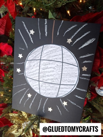 A handmade disco ball chalk art project on black paper, decorated with white chalk lines and gold star stickers, is displayed against a Christmas tree with red poinsettias. @GLUEDTOMYCRAFTS is written at the bottom.