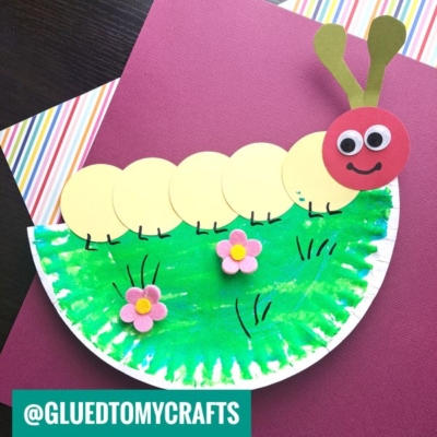 A Circle Punch Caterpillar Craft featuring a green-painted paper plate semi-circle with pink flowers and a caterpillar made of yellow circles, a red head, googly eyes, and brown antennae, displayed on colorful paper backgrounds.