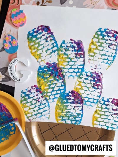 A white paper shows stamped, colorful fish scales made with a paint palette and brush. Decorative felt flowers and Easter egg cutouts are nearby. The handle @GLUEDTOMYCRAFTS is written at the bottom.