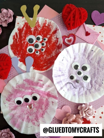 Three paper plate monsters, inspired by coffee filter crafts for kids, are decorated with paint, googly eyes, and paper cutouts. They’re displayed on a pink, heart-themed background with craft supplies and hearts. The @GLUEDTOMYCRAFTS watermark is at the bottom right.