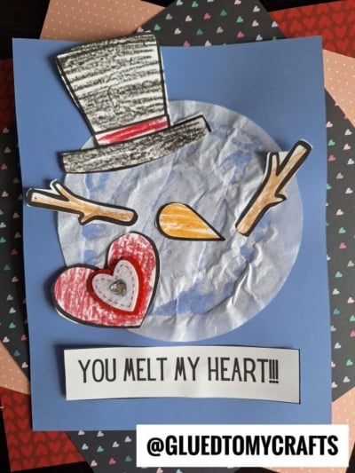 A handmade snowman craft on blue paper, featuring a crumpled white circle like those in coffee filter crafts for kids, paper hat, stick arms, heart shapes, and the message YOU MELT MY HEART!!! at the bottom with a @GLUEDTOMYCRAFTS tag.