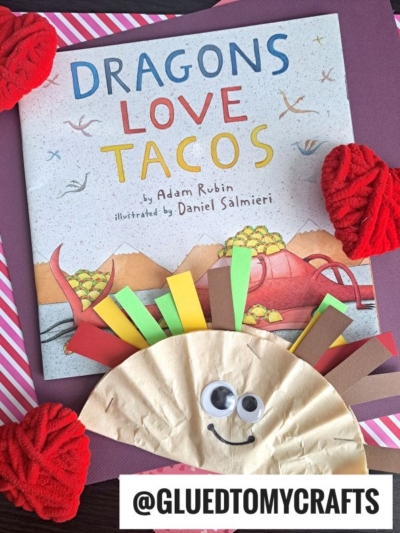 A childrens book titled Dragons Love Tacos is surrounded by red yarn pom-poms. Below the book is a taco craft, similar to coffee filter crafts for kids, made from a paper plate, colorful paper strips, and googly eyes. @GLUEDTOMYCRAFTS appears at the bottom.