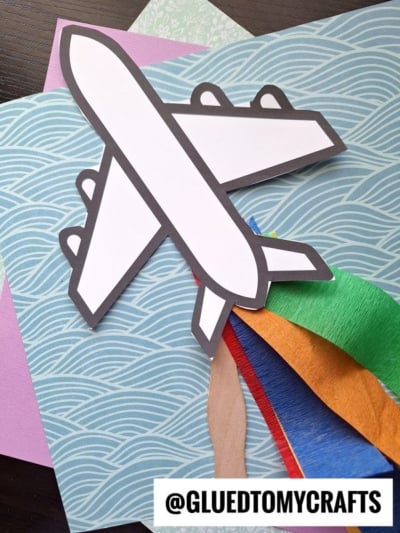 A paper airplane craft with a white body and black outline rests on patterned, colored paper and strips of tissue paper, perfect for those looking for creative crepe paper craft ideas. The text @GLUETOMYCRAFTS appears at the bottom.