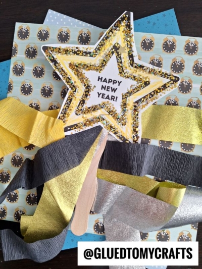 Assorted New Year’s craft supplies, including patterned papers, gold and silver star decorations reading Happy New Year!, yellow and gray crepe paper for creative crepe paper craft ideas, craft sticks, and a @gluedtomycrafts label.