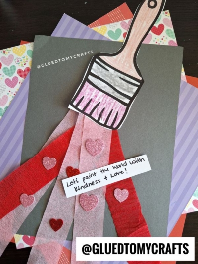 A charming crepe paper craft idea featuring a paper paintbrush with red and pink crepe paper paint, glitter hearts, and a note saying "Let's paint the hand with kindness & love!" on layered colorful paper backgrounds.