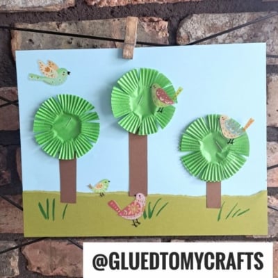 A children’s craft shows three trees made from green cupcake liners and brown paper trunks on a paper landscape, with colorful birds drawn on and around the trees—perfect inspiration for cupcake liner card crafts. “@GLUEDTOMYCRAFTS” appears at the bottom.