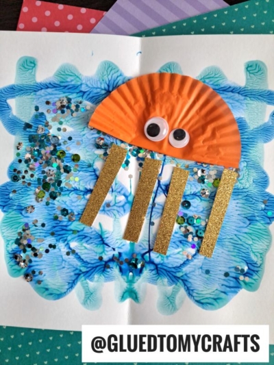 A colorful jellyfish PAINT SPLAT CRAFT made with an orange cupcake liner, googly eyes, gold glitter tentacles, and a blue paint background with scattered sequins. @GLUEDTOMYCRAFTS is written at the bottom.