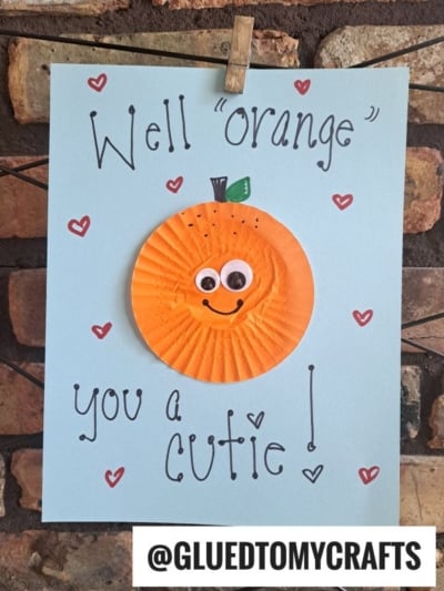 A handmade cupcake liner card crafts a sweet message: "Well orange you a cutie!" An orange cupcake liner smiles with googly eyes and a green leaf, surrounded by red hearts. @GLUEDTOMYCRAFTS is at the bottom.