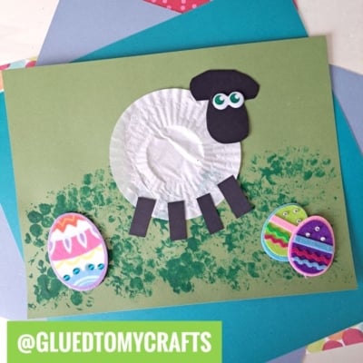 A fun children’s cupcake liner card craft featuring a sheep made from a white cupcake liner and black paper on a green background, with colorful decorated Easter eggs and a painted green grass effect. @GLUEDTOMYCRAFTS is written at the bottom.