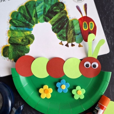 A paper plate craft of a caterpillar with red and green circles, googly eyes, and felt flowers sits beside the book The Very Hungry Caterpillar by Eric Carle. @GLUEDTOMYCRAFTS is printed at the bottom.