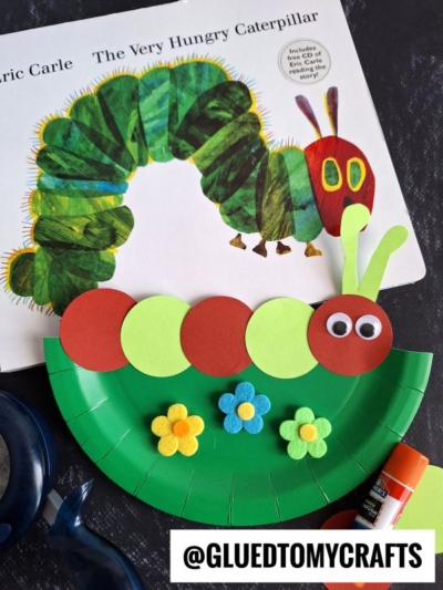 A paper plate craft of a caterpillar with red and green circles, googly eyes, and felt flowers sits beside the book The Very Hungry Caterpillar by Eric Carle. @GLUEDTOMYCRAFTS is printed at the bottom.