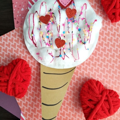 A handmade paper ice cream cone craft with a white cotton ball scoop, decorated with red hearts, colored sprinkles, and pink yarn, sits on layered patterned paper with red yarn hearts and the text @GLUEDTOMYCRAFTS at the bottom.