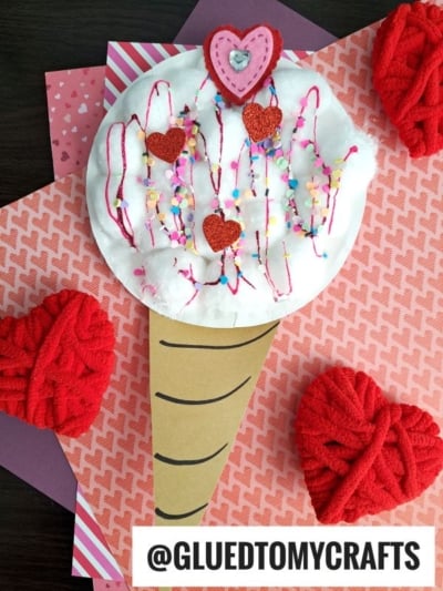 A handmade paper ice cream cone craft with a white cotton ball scoop, decorated with red hearts, colored sprinkles, and pink yarn, sits on layered patterned paper with red yarn hearts and the text @GLUEDTOMYCRAFTS at the bottom.