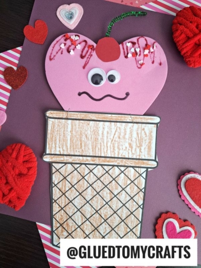 A handmade ice cream craft with a pink heart-shaped scoop, googly eyes, a cherry on top, and a crayon-colored cone—perfect for heart shaped crafts or valentine art project ideas. Surrounded by hearts with @GLUEDTOMYCRAFTS at the bottom.