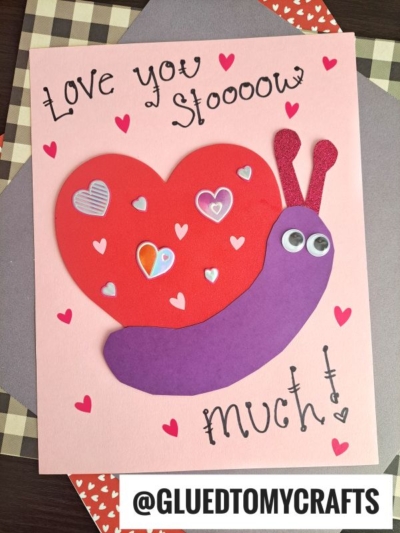 A handmade Valentine’s card, perfect for valentine art project ideas, features a snail with a red heart shell, purple body, googly eyes, and heart stickers. Text reads Love you sloooow much! on a pink background.