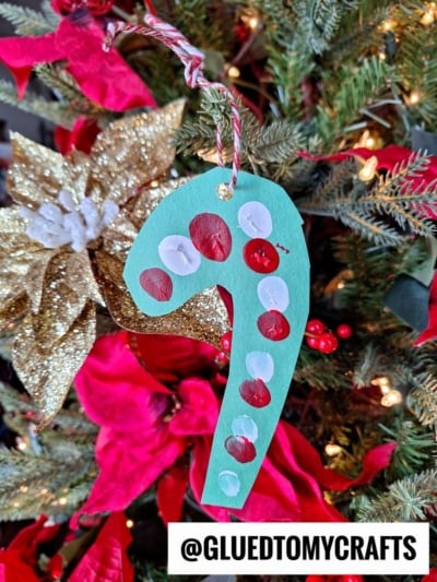 A handmade green paper candy cane ornament with red and white dots hangs on a Christmas tree decorated with red poinsettias and a gold glittery flower. Text at the bottom reads @GLUEDTOMYCRAFTS.