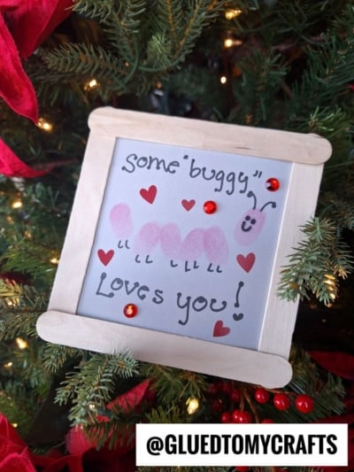 A framed drawing on a Christmas tree shows a caterpillar made from pink fingerprints with the words Some buggy loves you! decorated with red heart sequins. The handle @GLUEDTOMYCRAFTS appears at the bottom.