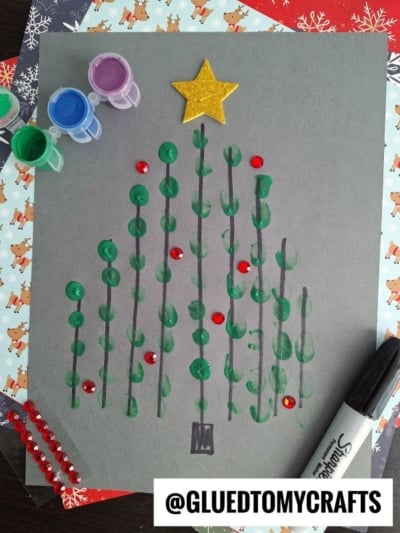 A Christmas tree made from green painted dots on gray paper, topped with a yellow star. Red gem stickers act as ornaments. Art supplies, a Sharpie, and the handle @GLUEDTOMYCRAFTS are visible around the artwork.