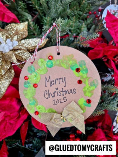 A round, brown ornament decorated with a green painted wreath, red sequins, and a gold bow reads Merry Christmas 2025. It hangs on a Christmas tree with red poinsettias and gold ribbon. Text: @GLUEDTOMYCRAFTS.