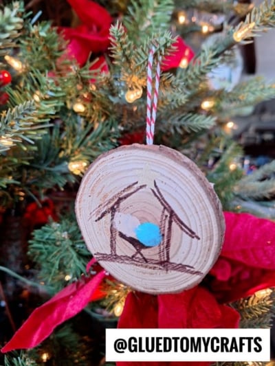 A round wooden ornament with a hand-drawn Nativity scene hangs on a Christmas tree with red ribbon decorations. The ornament has a striped string and features a simple depiction of Mary, Joseph, and baby Jesus.