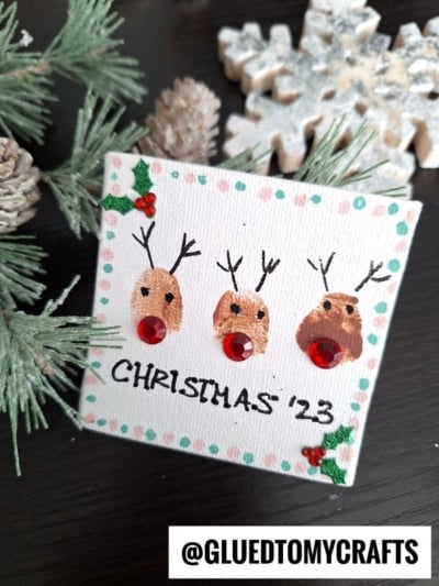 A small canvas features three thumbprint reindeer with red gem noses, antlers, and holly decorations. CHRISTMAS 23 is written below. Pine branches, pinecones, and a snowflake ornament are in the background.