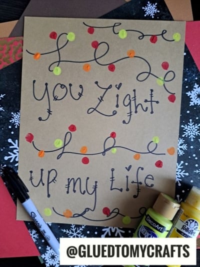 A decorated brown card reads You light up my life in cursive, with colorful dots resembling Christmas lights. Art supplies, including paint bottles, a marker, and craft paper, are arranged around the card.