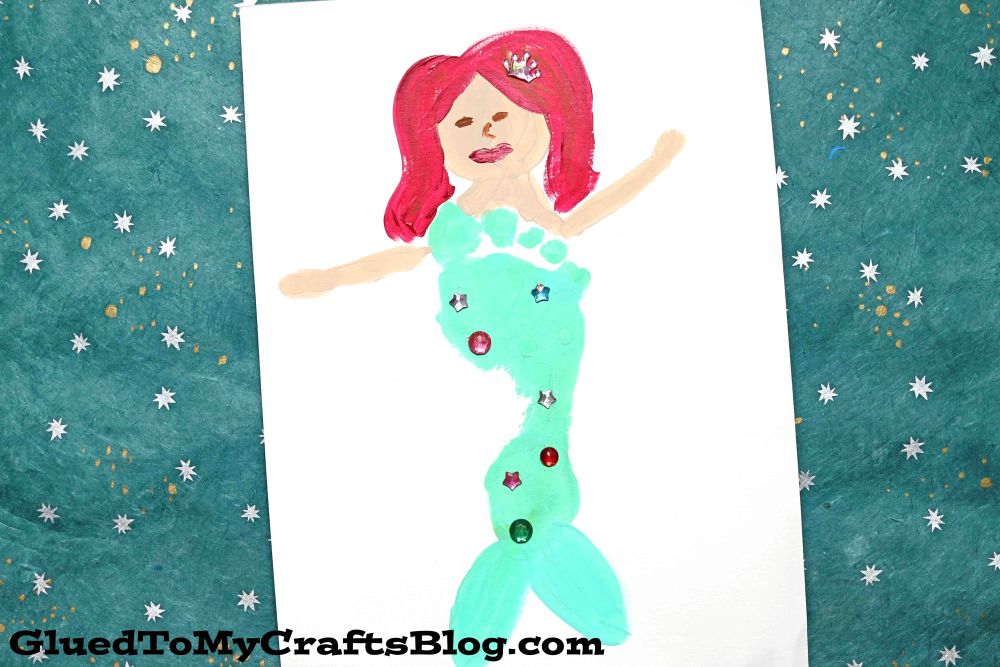 A childs painting of a mermaid with red hair, a crown, and a green tail decorated with colorful sequins, on white paper atop a teal, star-patterned background.
