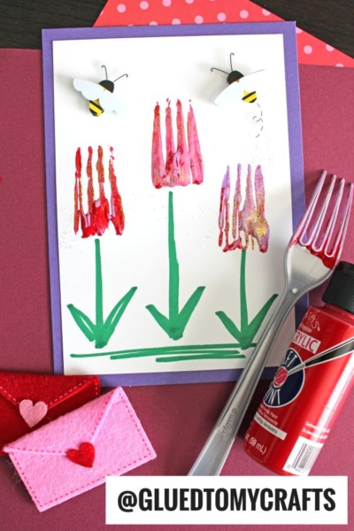 A handmade card features fork painted tulips with green stems and leaves, crafted using a fork and red paint. Accents include a felt envelope, paint bottle, and paper bees on purple paper; @GLUEDTOMYCRAFTS text is visible.