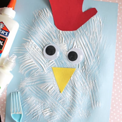 A childrens chicken craft made with a blue paper, white paint strokes, large googly eyes, a yellow triangle beak, and a red paper handprint for the comb. A plastic fork and glue bottle are next to the craft.