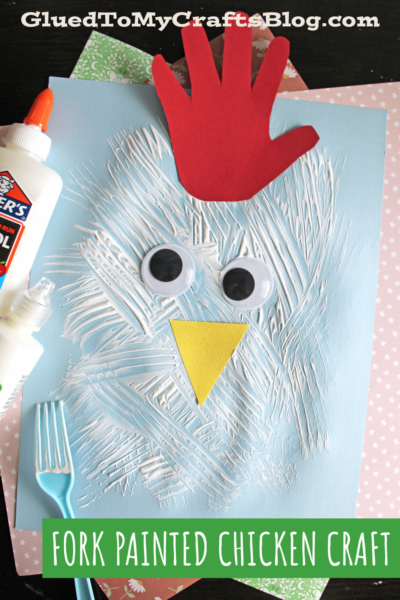A childrens chicken craft made with a blue paper, white paint strokes, large googly eyes, a yellow triangle beak, and a red paper handprint for the comb. A plastic fork and glue bottle are next to the craft.