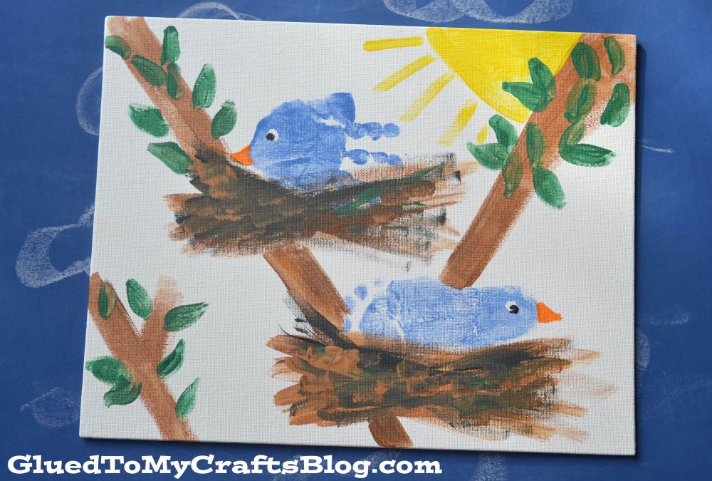 A child’s painting of two blue birds with orange beaks sitting in brown nests on tree branches. Green leaves and a yellow sun are visible in the background. The image has a blue background and a blog watermark.
