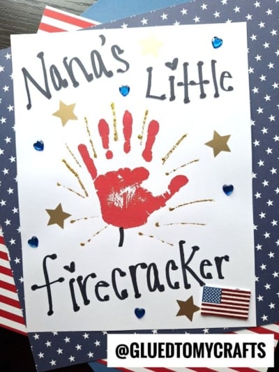 A white card reads Nanas Little Firecracker with a red handprint in the center, gold stars, blue heart gems, and a small American flag sticker—perfect for 4th of July keepsakes. The card sits on star-patterned paper. @GLUEDTOMYCRAFTS is at the bottom.