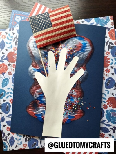 A handmade 4th of July keepsake featuring a cutout of a white hand holding a small American flag, set on colorful patriotic paper with glitter and a floral backdrop. @GLUEDTOMYCRAFTS is written at the bottom.