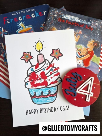 A handmade card with a cupcake and “Happy Birthday USA!” sits beside 4th of July keepsakes—a red badge and two patriotic-themed children’s books—each adorned with festive red, white, and blue decorations.