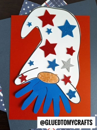 A handmade gnome craft on red paper features a white hat decorated with red, blue, and silver stars, a tan oval nose, and a blue handprint-shaped beard—perfect for 4th of July keepsakes with patriotic patterned papers layered beneath.