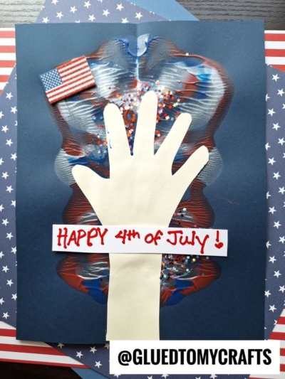 A handprint cutout is glued to dark paper, decorated with red, white, and blue paint and glitter. A small US flag sits above, "Happy 4th of July!" is written in red—a perfect 4th of July keepsake. @GluedToMyCrafts at the bottom.