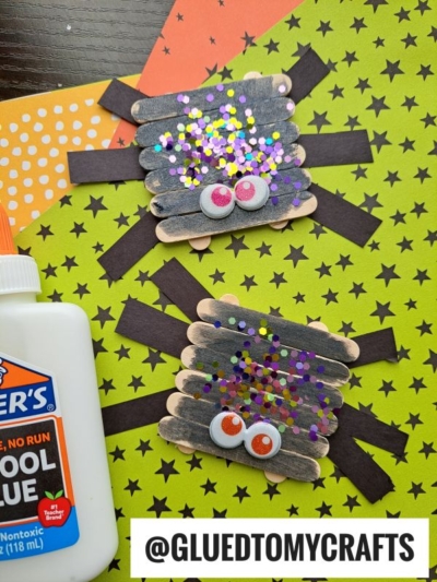 Two spider crafts made from black-painted popsicle sticks, googly eyes, and colorful glitter sit on green and orange patterned paper. Perfect for INSECT CRAFT IDEAS, a bottle of glue is visible and @GLUEDTOMYCRAFTS is written at the bottom.