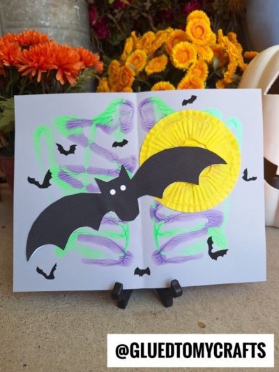 A handmade Halloween card features a large black bat and yellow moon, with vibrant PAINT SPLAT CRAFTS in purple and green, small bat cutouts, and orange flowers in the background. @GLUEDTOMYCRAFTS is displayed at the bottom.