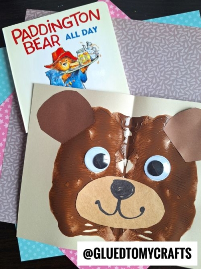 A Paddington Bear book and a cute bear face made from brown paint splat crafts, googly eyes, and paper ears and nose are displayed on colorful papers. The Instagram handle @gluedtomycrafts is at the bottom.