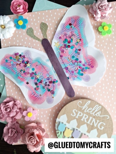 A colorful butterfly craft made with marker lines, confetti, and a wooden stick for the body, surrounded by pink and white flowers on a pastel polka-dot background with a “hello SPRING” sign—perfect inspiration for PAINT SPLAT CRAFTS.